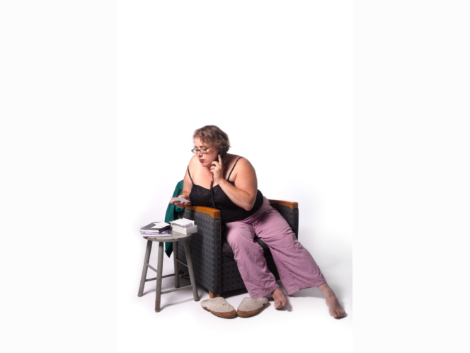 A white woman in glasses, pajamas, and a tank-top, sits in a chair, looking through cards and holding a phone to her ear.