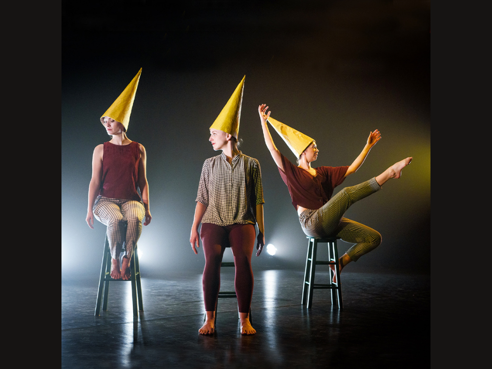 Three dancers are sitting on green stools with yellow dunce caps on. Two are looking away from the dancer to the right who has one leg extended.