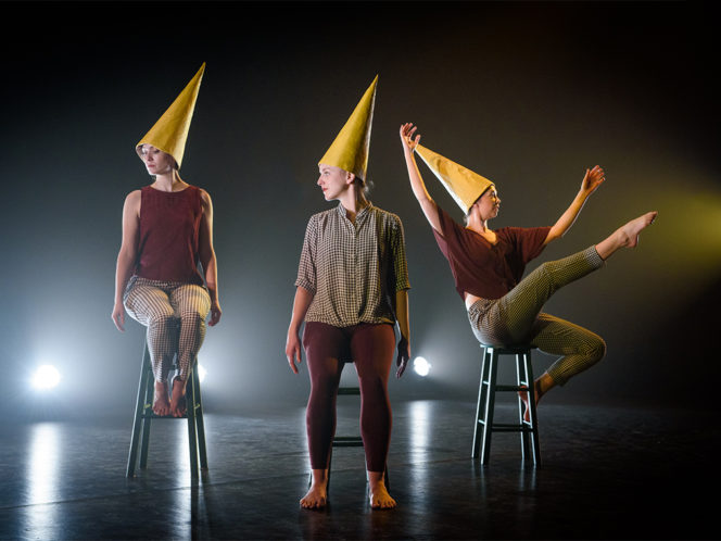 Three dancers are sitting on green stools with yellow dunce caps on. Two are looking away from the dancer to the right who has one leg extended