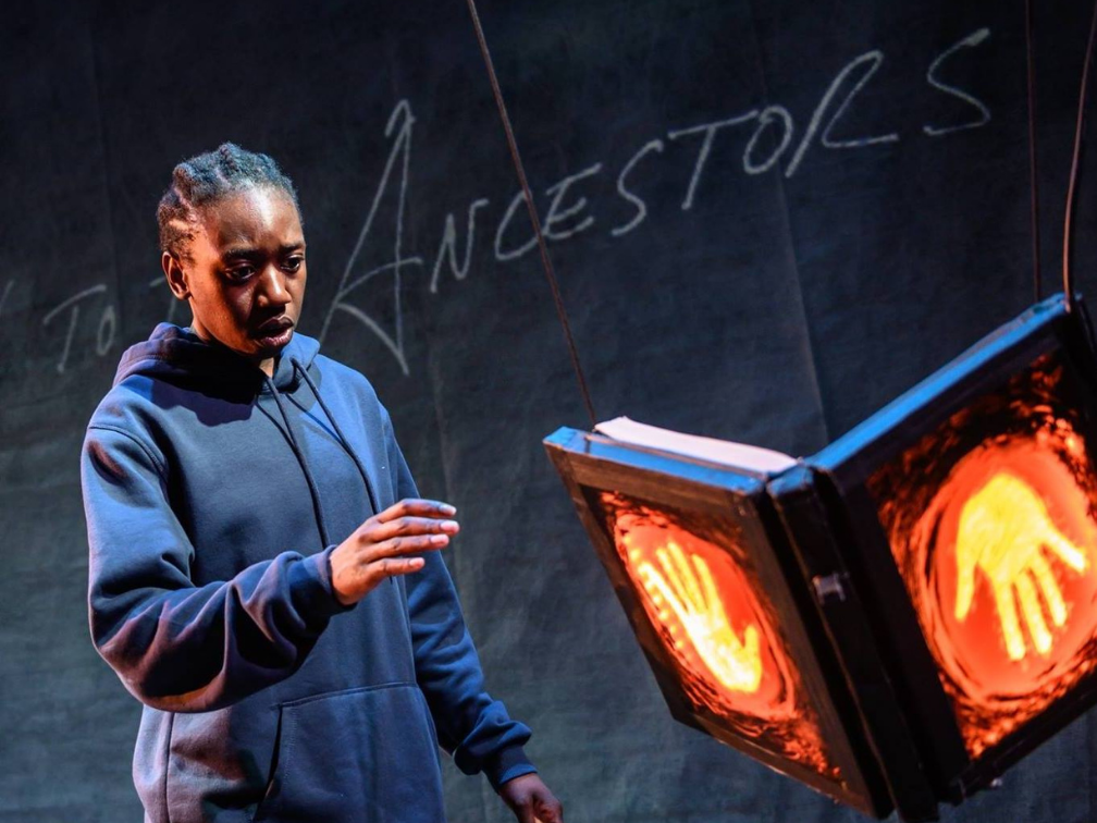 The actor, Makambe is wearing a blue hoodie, reaching towards a light up sign. The back wall has chalkw riting that reads The Ancestors.