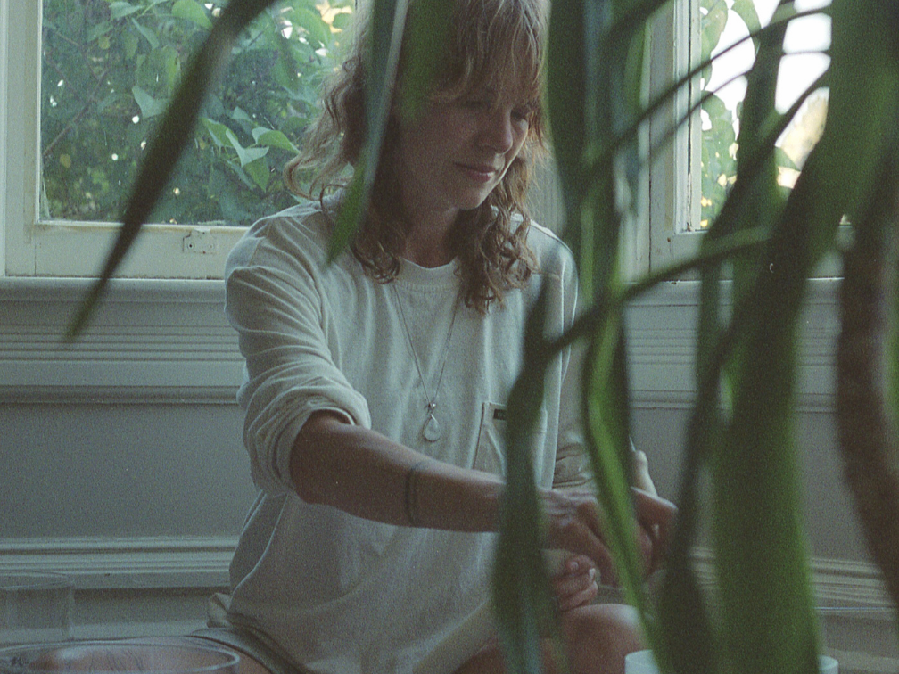 Charlie wears a cream shirt, sitting against a window surround by plants, playing a bowl.