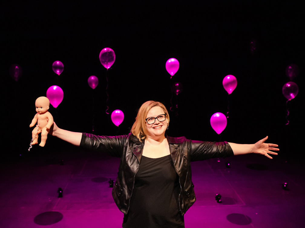 Jamie stands on stage, arms outstretched, holding a baby doll. Behind ehr are several ballons on strings, lit bright pink.