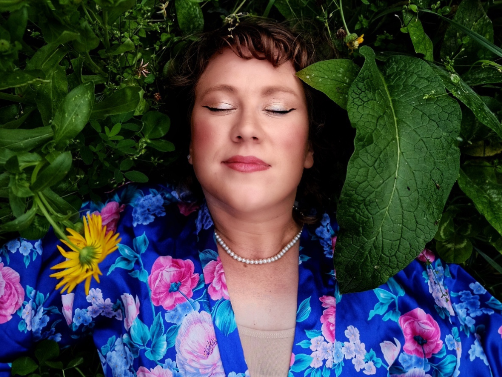 A white-skinned woman lies in a bed of deep green foliage, her eyes closed in an expression of quiet ecstasy. She wears a low-cut blue silk floral robe and a string of pearls around her neck.