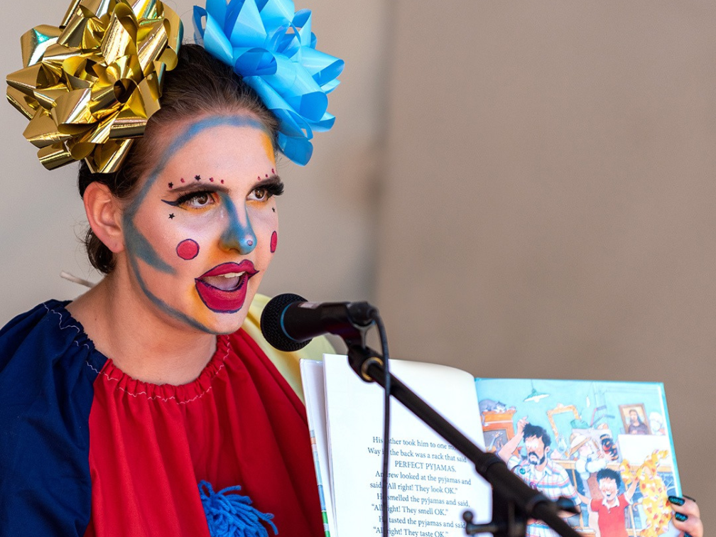A drag performer in bright makeup with big bows on tehir end sits behind a microphone, reading a storybook