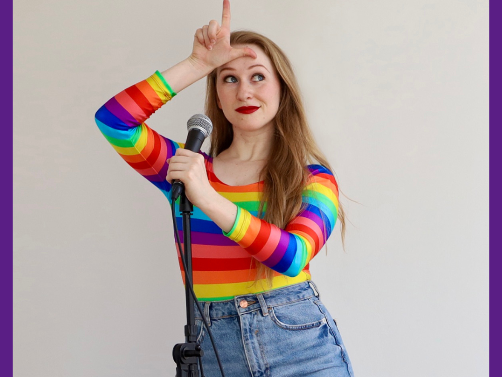 A blonde girl with a cheeky smile, a long-sleeve rainbow body suit and light-washed ripped jeans, stands centre in the frame behind a microphone, holding her right hand in the shape of an “L” on her forehead.