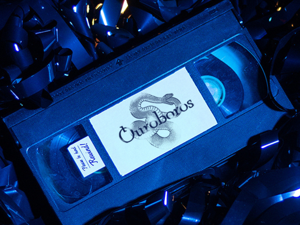 A VHS cassette sits on a pile of loose tape. The faded label has the word Ouroboros on top of a snake eating it’s own tail.