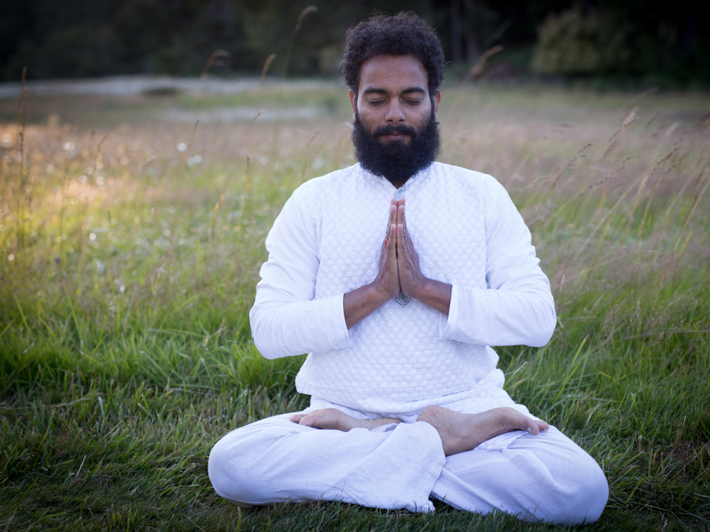 Amit sits on the grass with his legs crossed and his palms together and eyes closed. He wears all white.