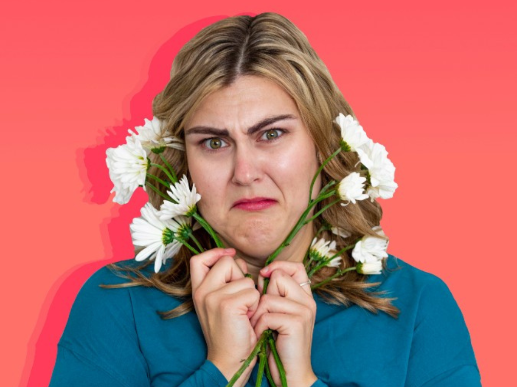 A white woman with blonde hair in a blue long sleeve shirt holding white daisies around her face in front of a coral red gradient background.