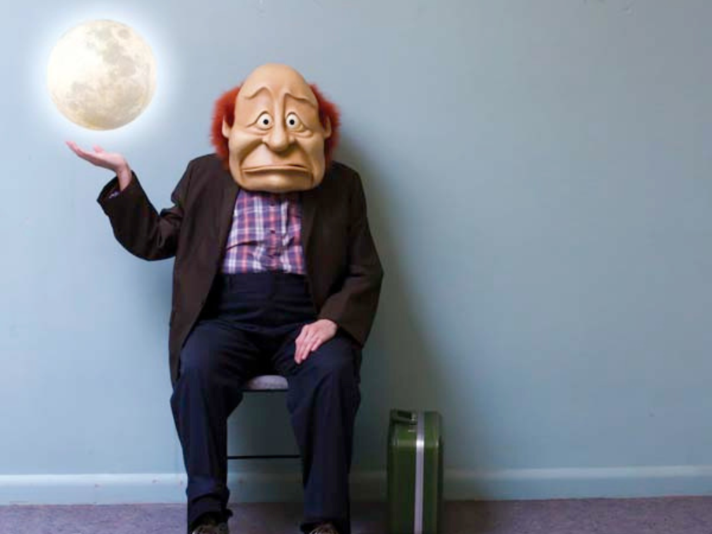 A character with a full sized face mask sits in a chair, holding a moon in an outstretched hand.