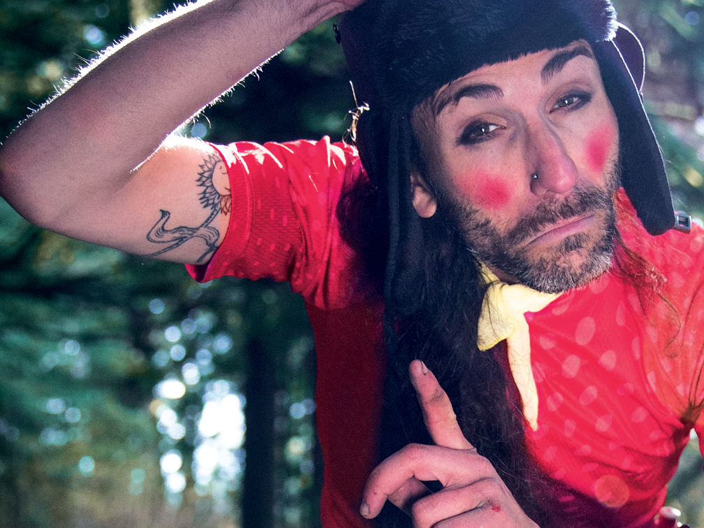 Stanczyk, a Polish clown, stands in a forest, with long brown hair and painted on black eyeshadow and rosey cheeks, wearing a black fur hat with ear flaps, a yellow neck scarf, and a red cycling shirt, holds his head, raises a finger, grimaces at the camera, whilst sitting on a just-visible bicycle.