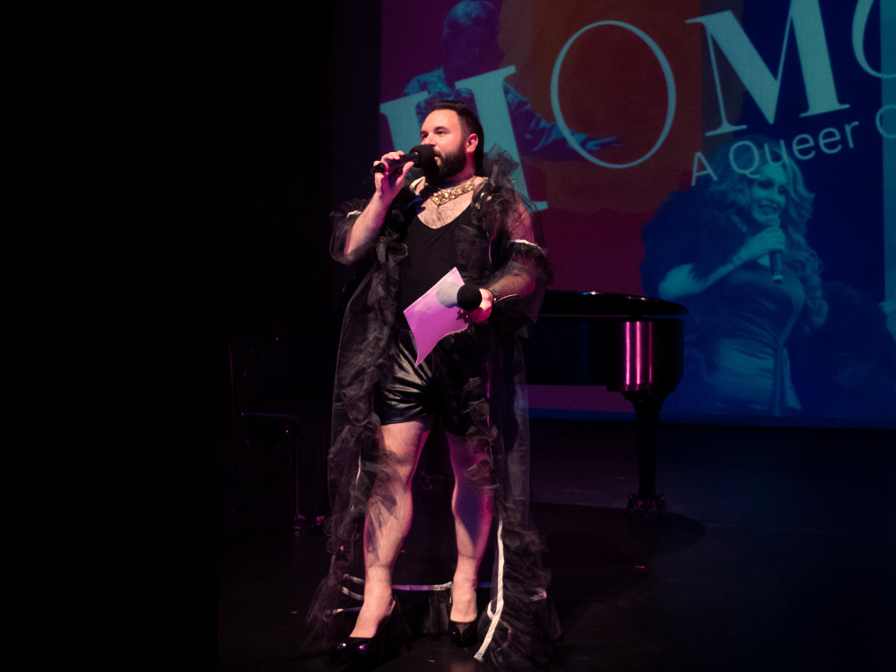 Sean stands on stage, holding a microphone and a paper. He is wearing black sorts, high heels and a black tulle cape. Behind im is a piano and a projection of the HOMO Cabaret poster.