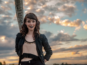 a person leans against a thin tree, they wear a dark coat and pants and a nude shirt with tassels on the bottom. The sun behind them is setting.