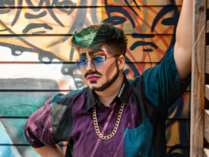 A person in drag leans against a wall. The mural behind them is colourful and abstract. They wear a purple and blue shirt, gold chain necklace and dramatic makeup. They have a beard and moustache and wear blue glasses.