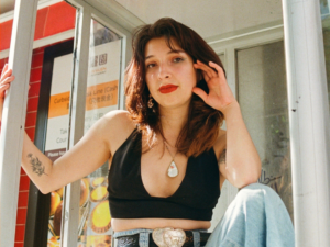 A person stands looking down at the camera in a phone booth. They wear red lipstick and holds their hand delicately beside their face.