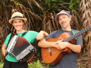 Two people stand, the one on the left leans their elbow on the others shoulder. They hold an accordion. The person to the right holds a guitar. Behind them are palm trees.