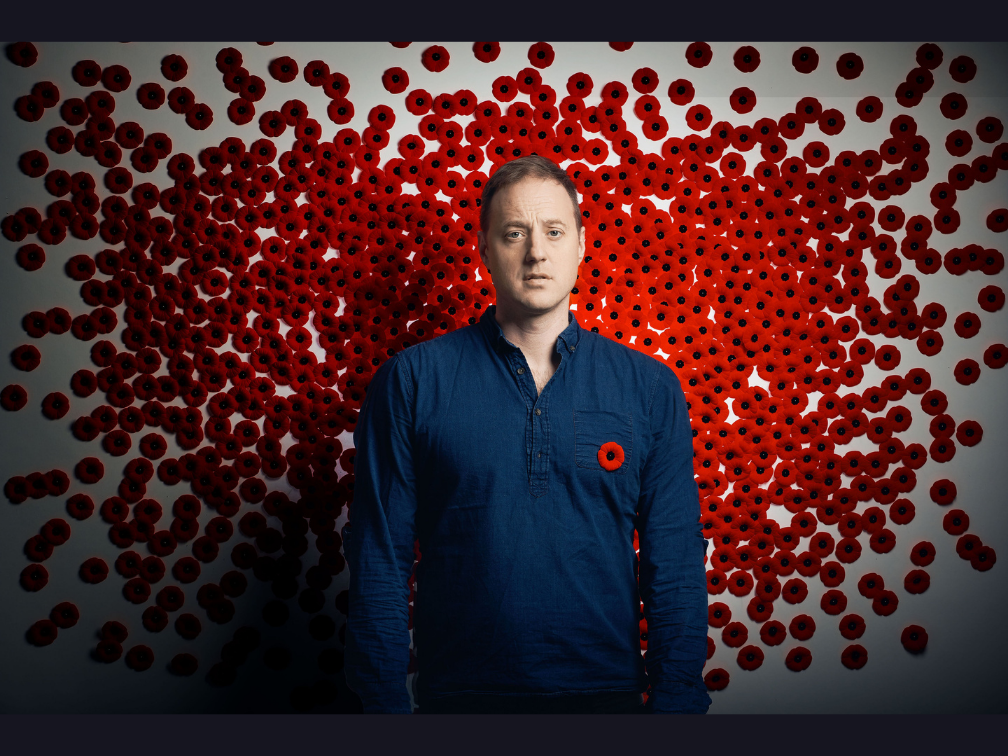 Brendan is wearing a blue shirt and a poppy, he is standing in front of a wall covered in red poppies.