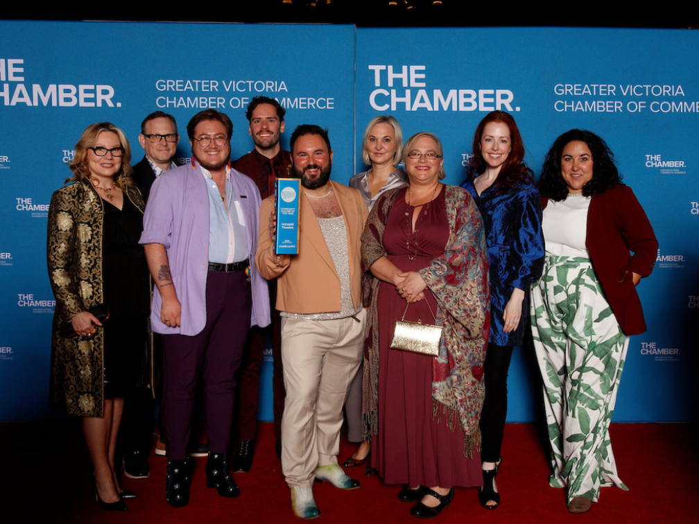 The INtrepid Theatre team stand on a red carpet in front of a blue backdrop, in the centre, Sean holds an award.