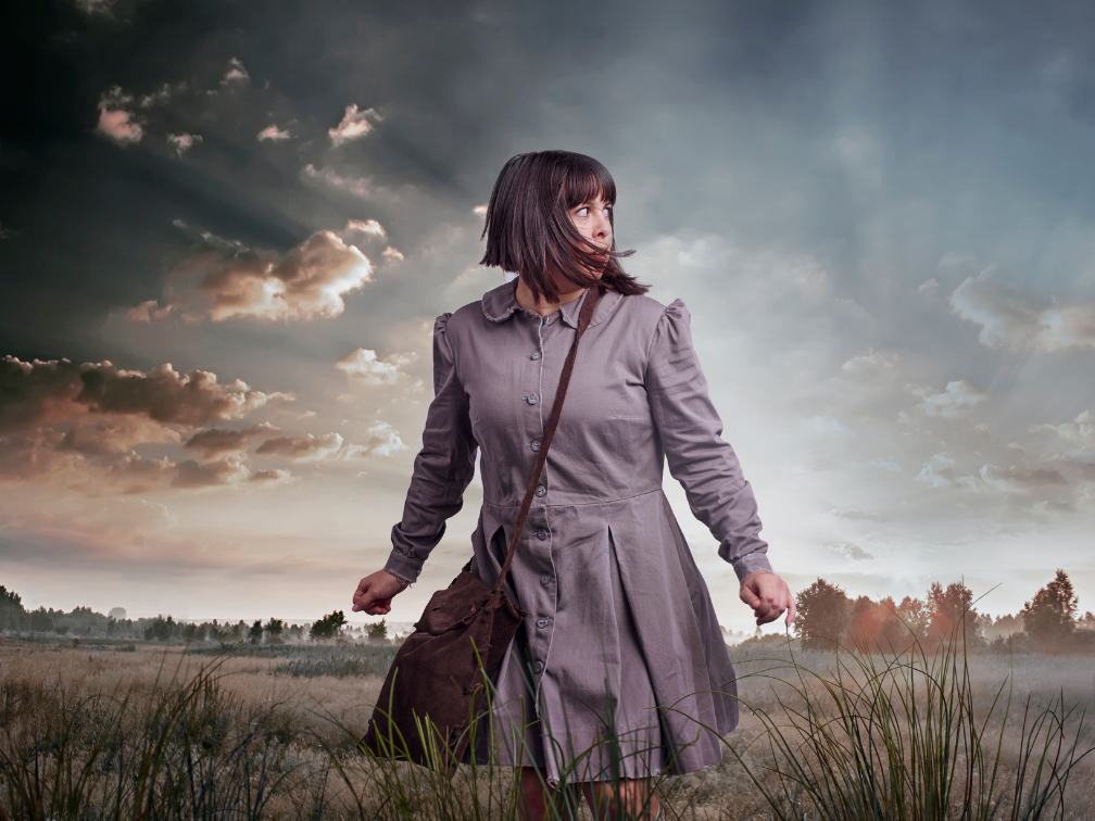 A woman in a grey school uniform stands amongst a field and an open sky.