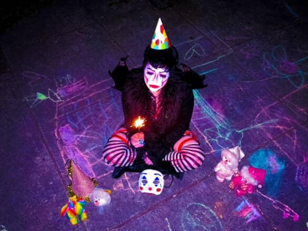 Drag Performer Phoenix HellFire sits on the concrete with a childrens birthday hat. They hold a sparkler and sit with clowns and a clown mask.