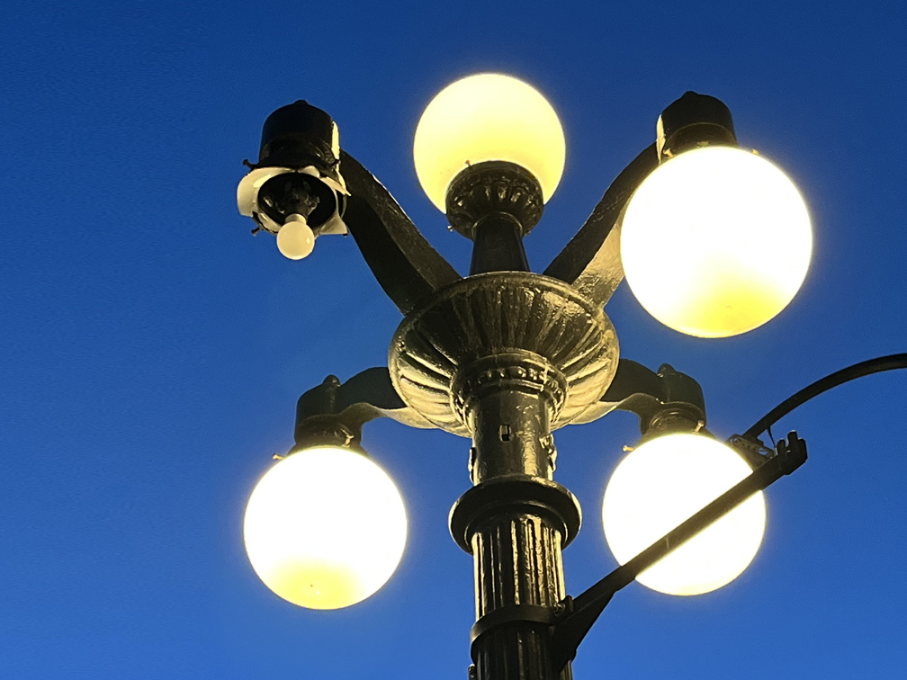 image shows a lit lamp-post over dark blue twilight sky with four hanging glass lights. one of the light covers is missing and is showing an exposed and unlit lightbulb.