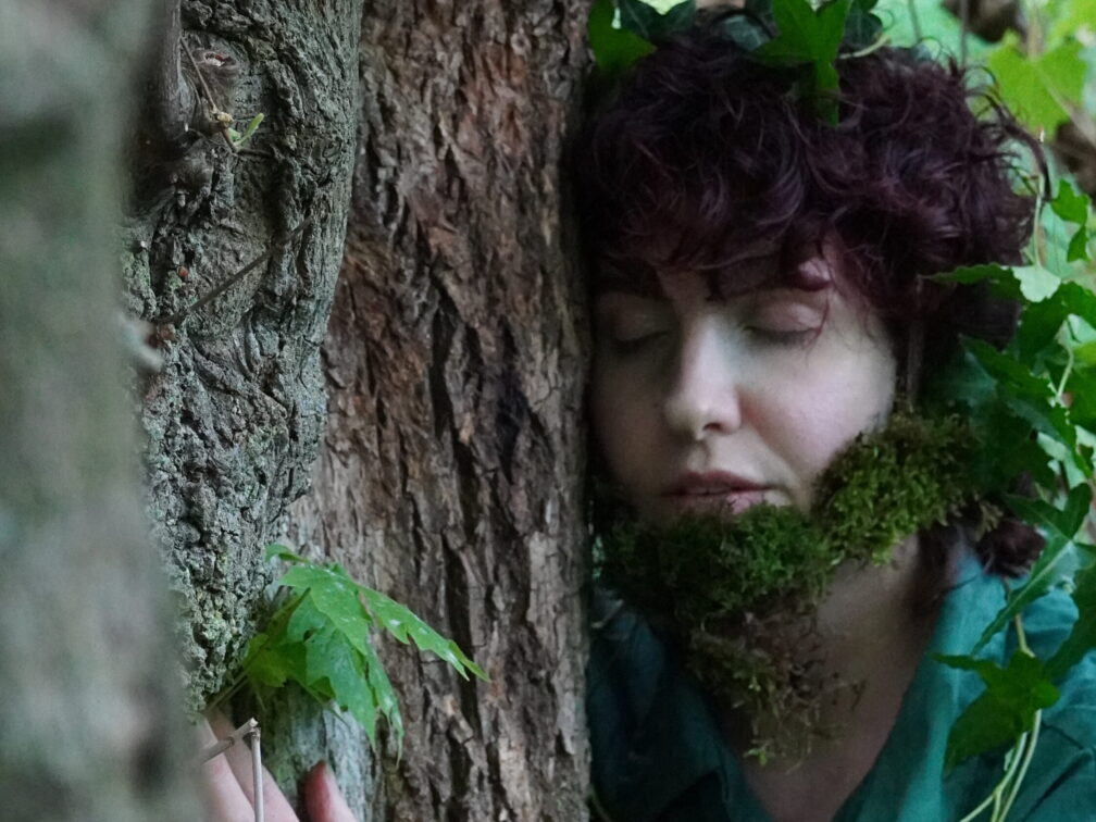 An actor leans on a Douglas Fir, peacefully connecting, with their hand placed lovingly on it's side. The actor is wearing a teal blue jumpsuit, and has moss covering their face in the shape of a beard.