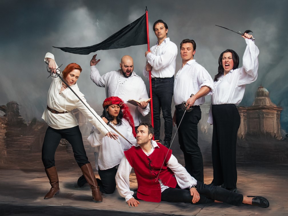 A tableau of Shakespearean actors: A man lays on the ground surrounded by two women and a man holding weapons. Another man shouts down at him. Another man holds a black waving flag. A final woman raises a sword and shouts.