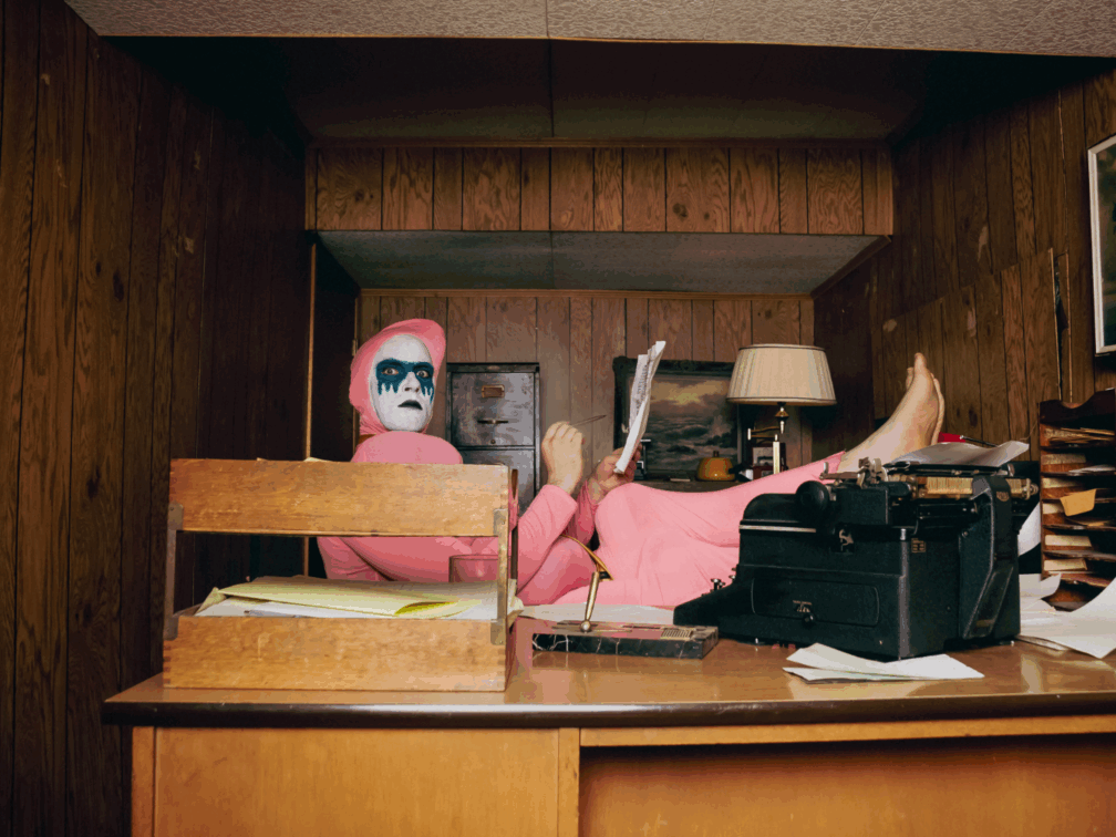 A man in a bulbous pink suit staring at the camera with his feet on the desk.