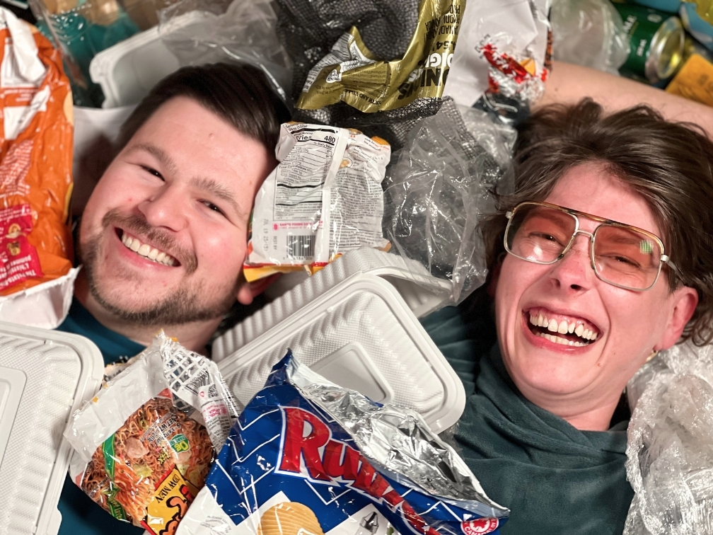 The faces of Logan Swain and Sadie Fox emerging from a pile of mixed recycling. Logan is smiling and Sadie is smirking.