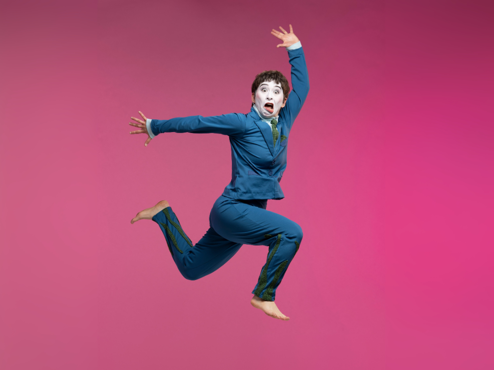 A performer in white mime makeup and a blue suite is jumping in the air, arms and legs extended, on a pink background.